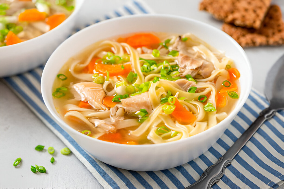 A bowl of chicken noodle soup with noodles, carrots, and green onions, served on a striped blue and white napkin.