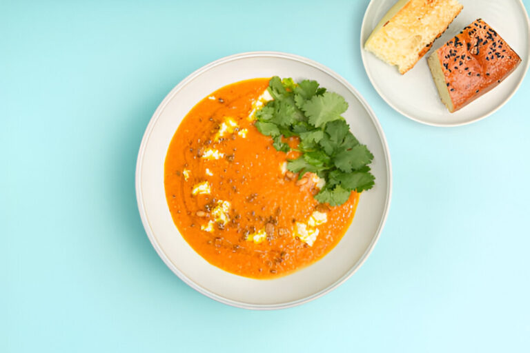 A bowl of vibrant orange soup garnished with fresh cilantro, next to a plate of bread rolls.