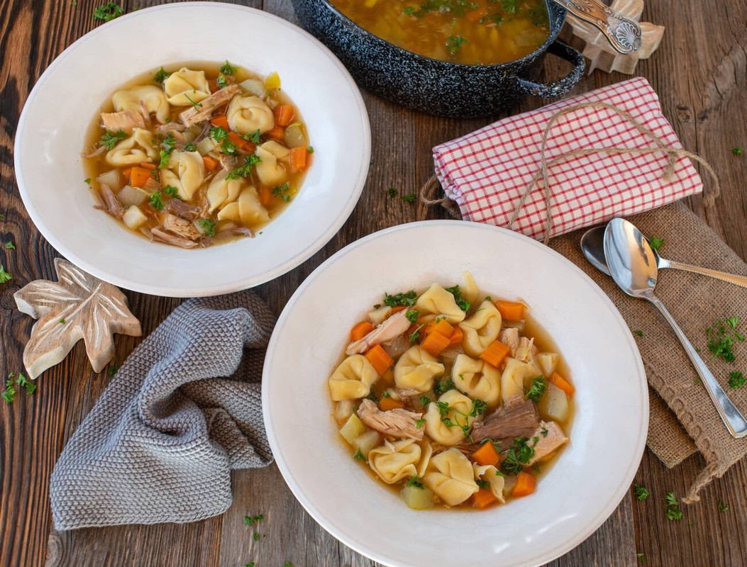Two bowls of homemade tortellini soup with carrots and chicken on a rustic wooden table.