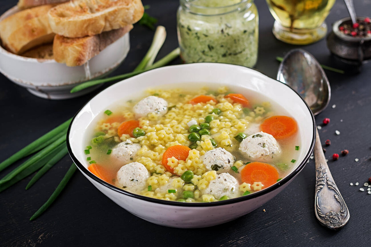 A bowl of soup with star-shaped pasta, meatballs, carrots, and green peas.