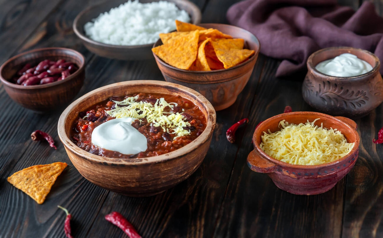 A delicious bowl of chili topped with shredded cheese and sour cream, surrounded by rice, tortilla chips, and bowls of kidney beans and shredded cheese.