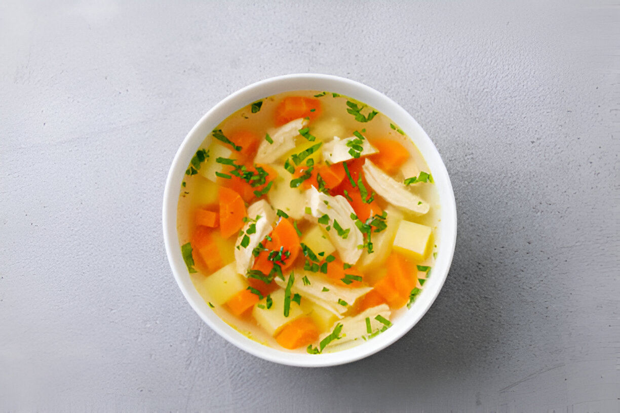 A bowl of chicken soup with vegetables including carrots and potatoes on a gray background.