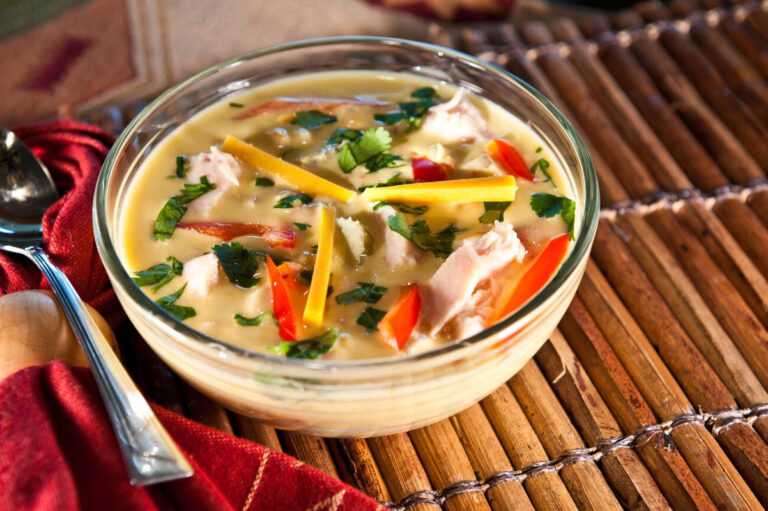 A warm bowl of creamy soup garnished with chicken, colorful peppers, and fresh cilantro, placed on a rustic wooden surface.