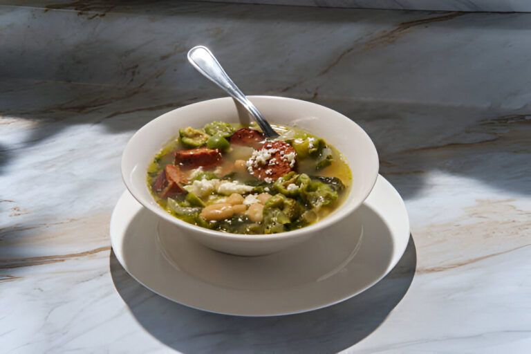 A bowl of green soup with vegetables and slices of sausage on a marble countertop.