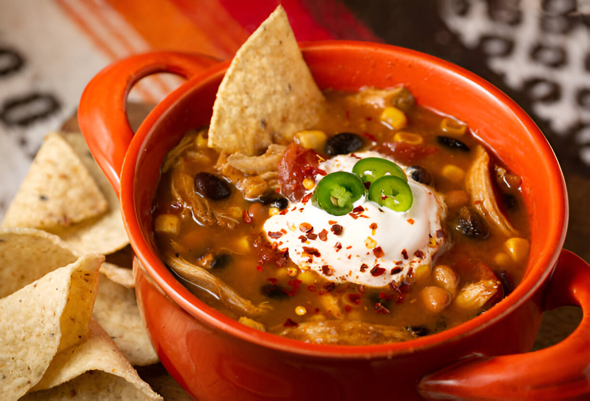 A vibrant orange bowl filled with chili topped with sour cream, jalapeños, and red pepper flakes, accompanied by tortilla chips.