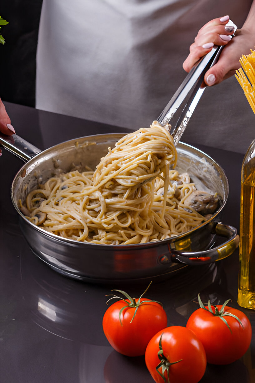 A person holding tongs with spaghetti in a pan, with fresh tomatoes and olive oil on the side.