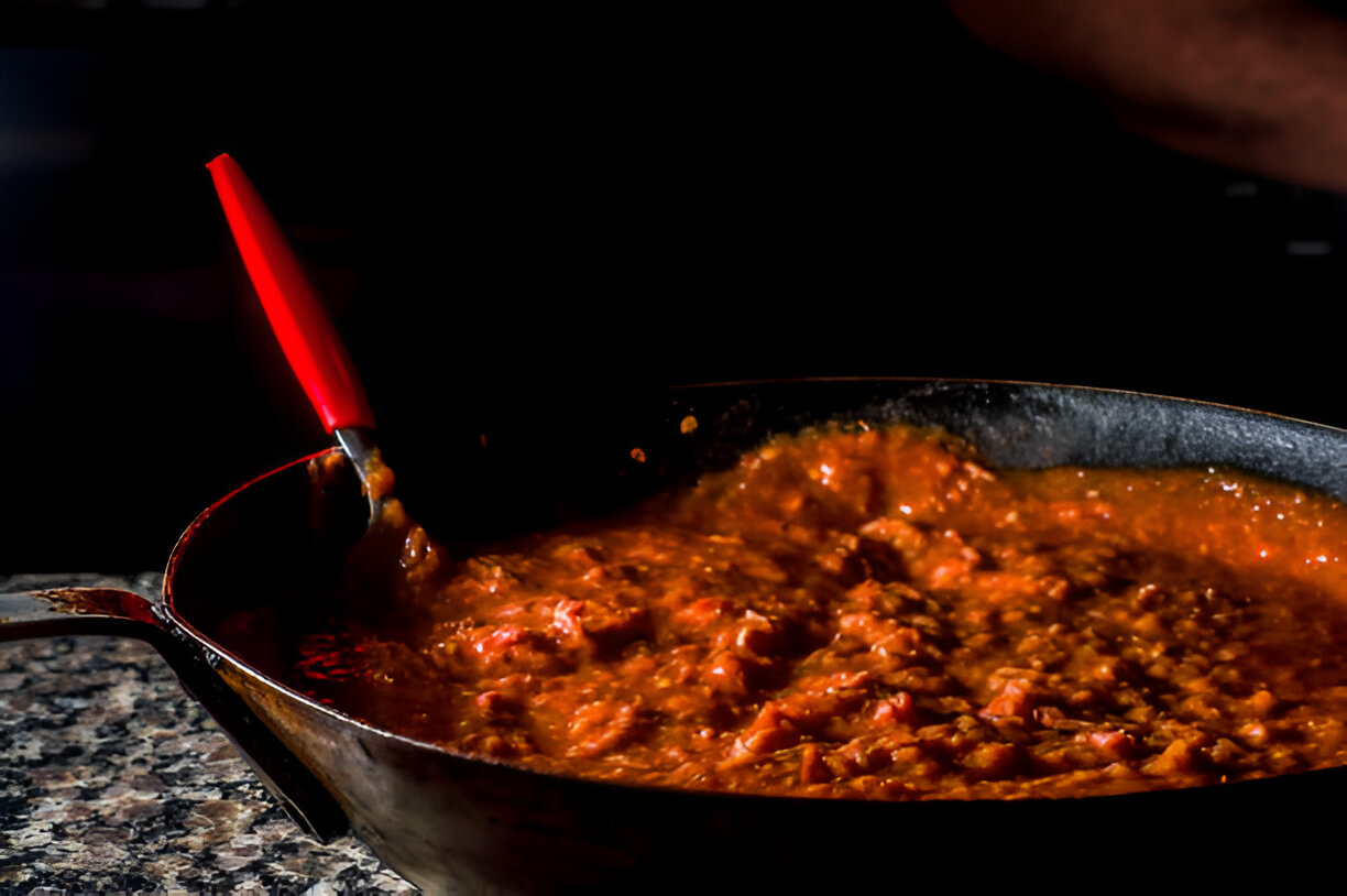 A pot of simmering sauce with a red-handled spoon stirring, set against a dark background.