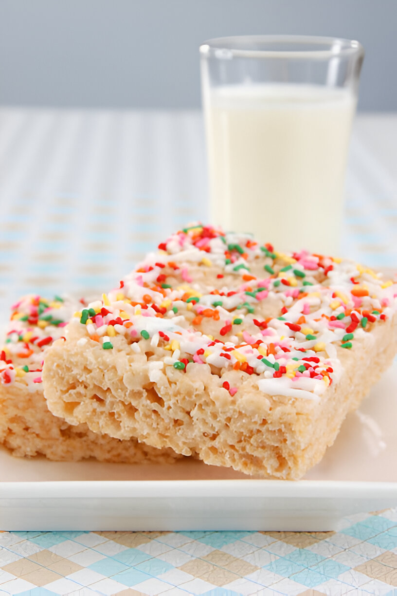 Two square pieces of Rice Krispies treats with colorful sprinkles on top, served on a white plate with a glass of milk in the background.
