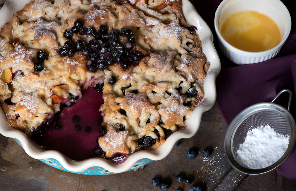 A freshly baked blueberry pie with a slice taken out, surrounded by blueberries and a small bowl of melted butter.