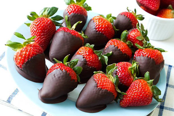 A plate of chocolate-covered strawberries arranged neatly, with some fresh strawberries in a bowl in the background.