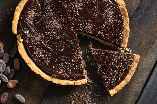 A chocolate tart with a slice removed, set on a wooden surface with cocoa beans nearby.
