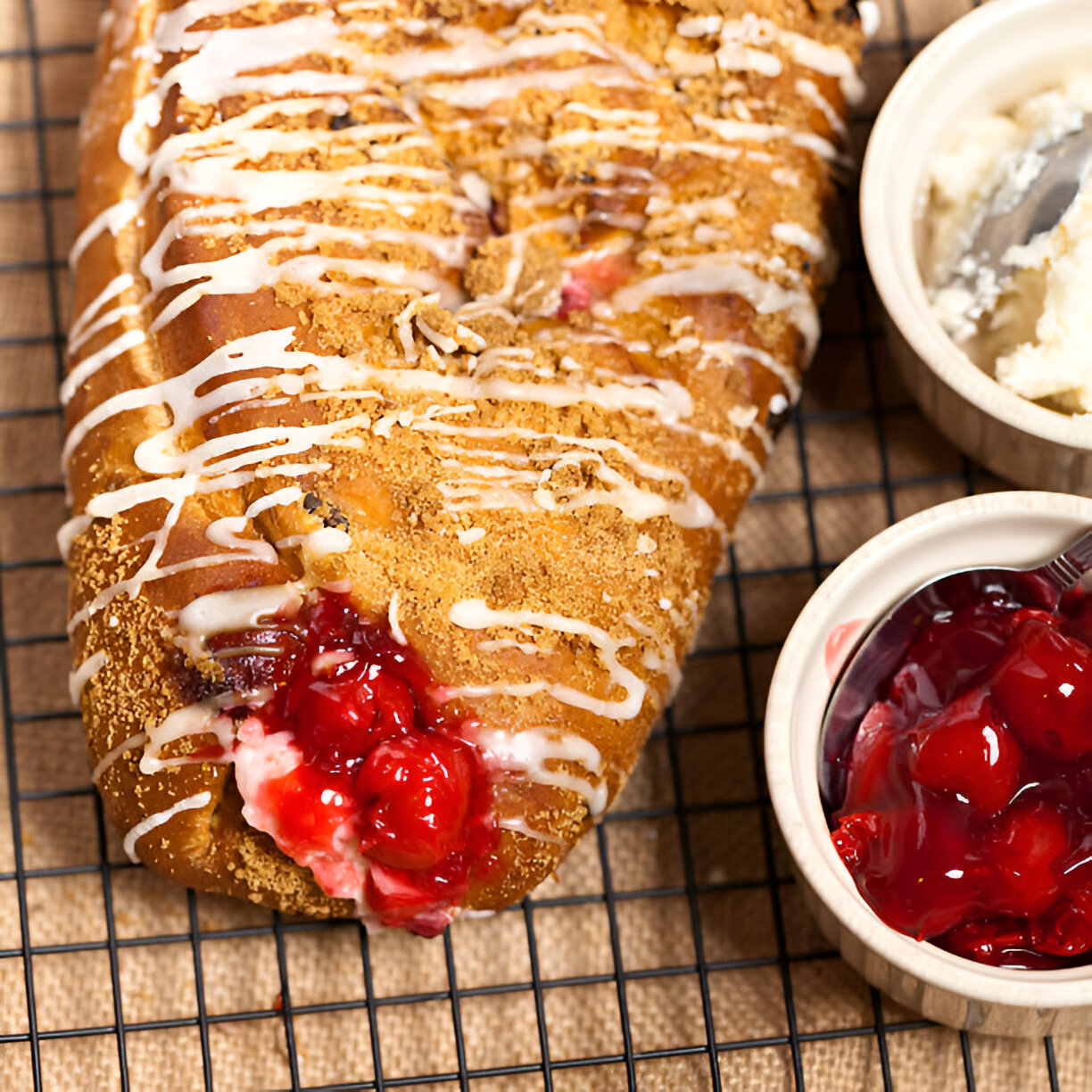 A close-up of a sweet pastry topped with icing and streusel, filled with red cherry filling, alongside a bowl of cherry topping and whipped cream.