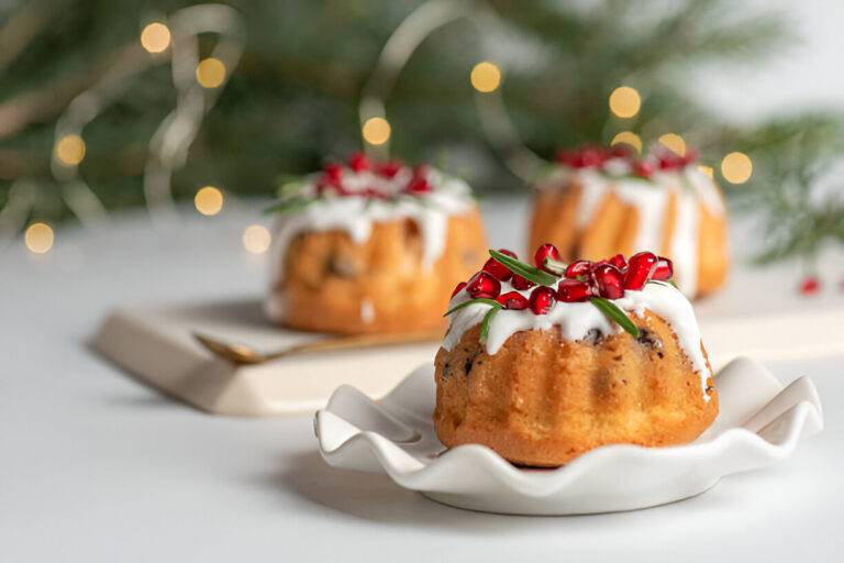 Three mini bundt cakes decorated with white icing, pomegranate seeds, and rosemary sprigs on a white plate against a blurred festive background.