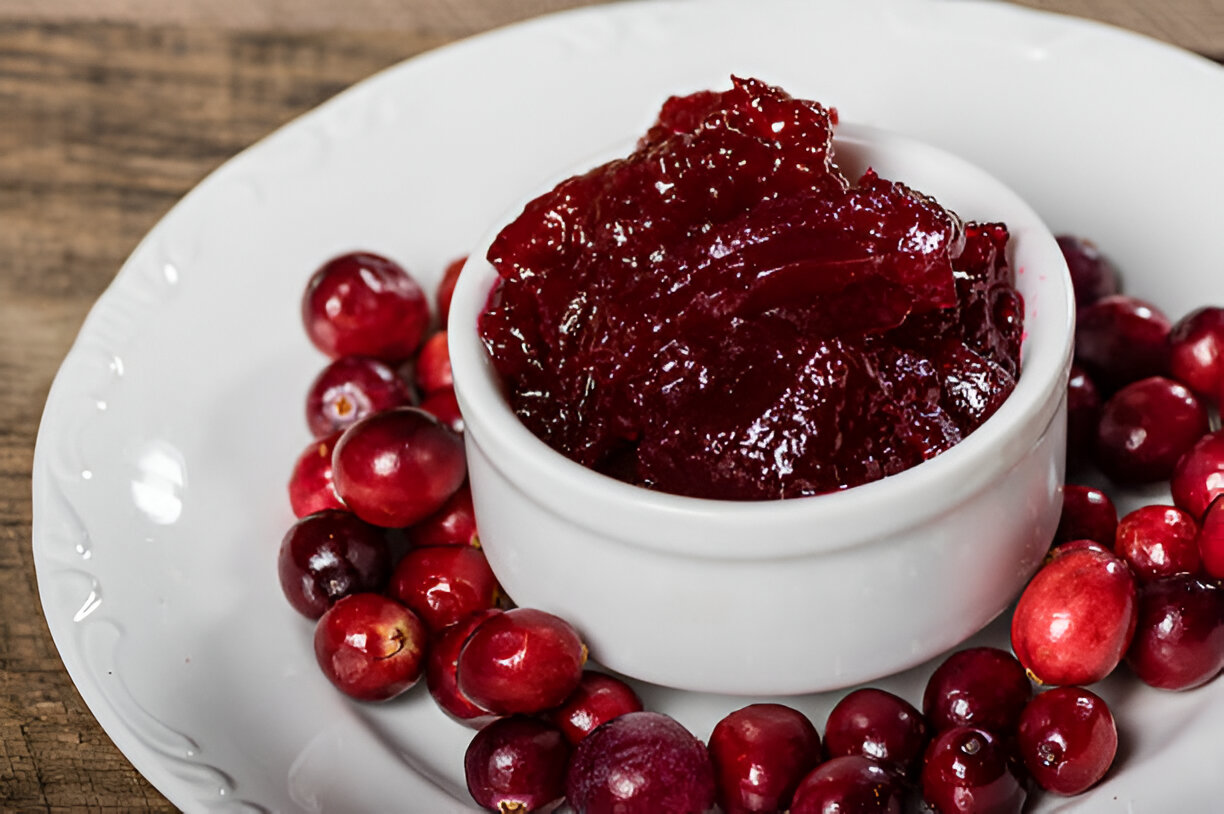 A white bowl filled with cranberry jelly surrounded by fresh cranberries on a white plate.