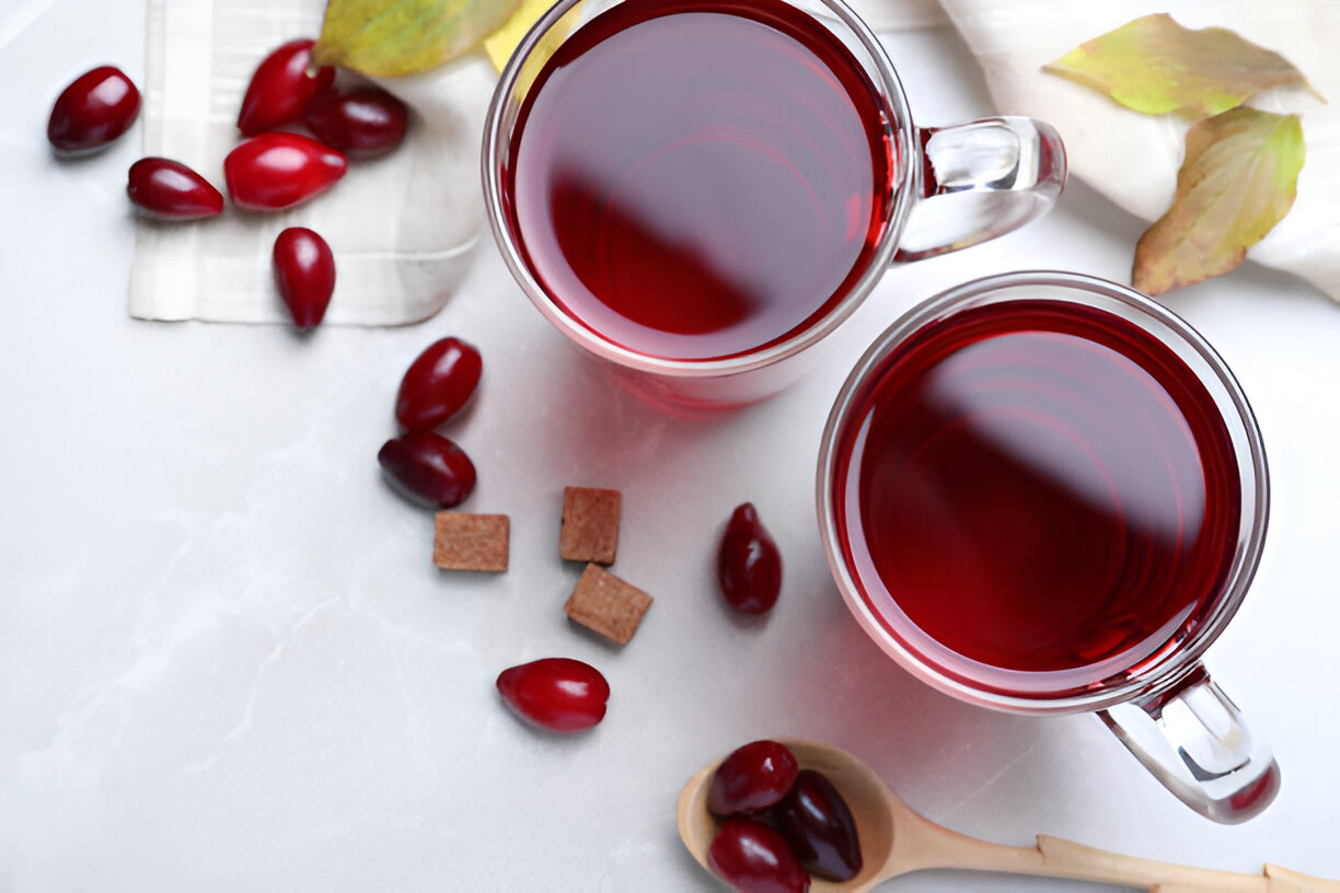 Two clear glass cups filled with deep red liquid, surrounded by red berries and brown sugar cubes on a light surface.