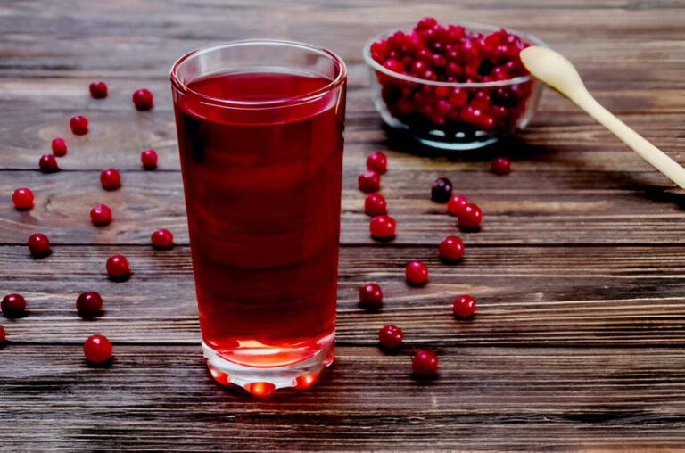 A glass of red beverage on a wooden table surrounded by fresh cranberries.