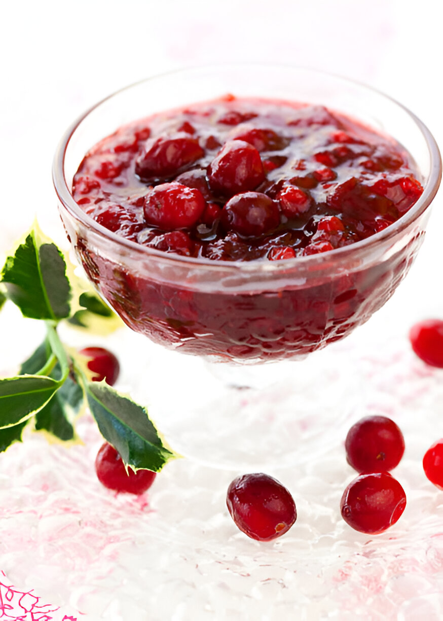 A glass dish filled with cranberry sauce, surrounded by fresh cranberries and green leaves.