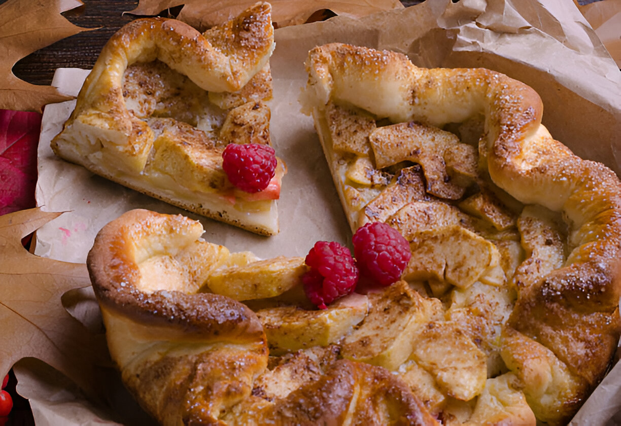 A close-up of a freshly baked apple pastry topped with raspberries on parchment paper, surrounded by autumn leaves.