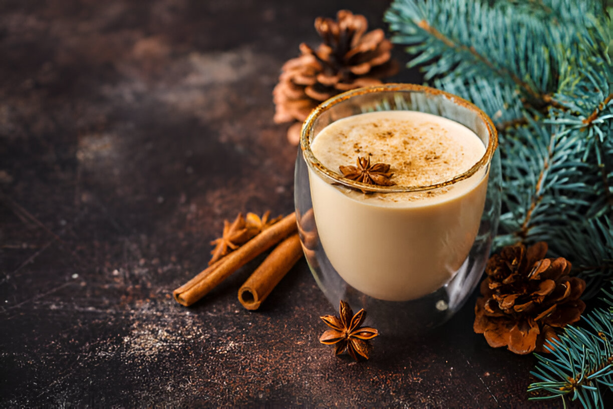 A glass of creamy drink topped with spices, surrounded by cinnamon sticks, pine cones, and evergreen branches.
