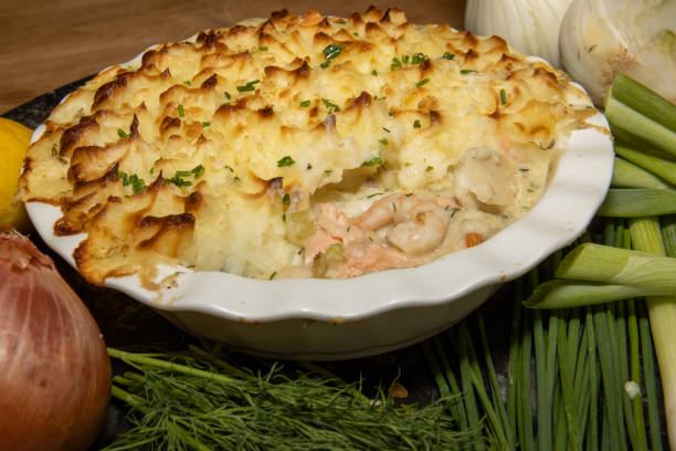 A delicious homemade seafood shepherd's pie topped with crispy golden mashed potatoes, surrounded by fresh vegetables including onions, leeks, and herbs.