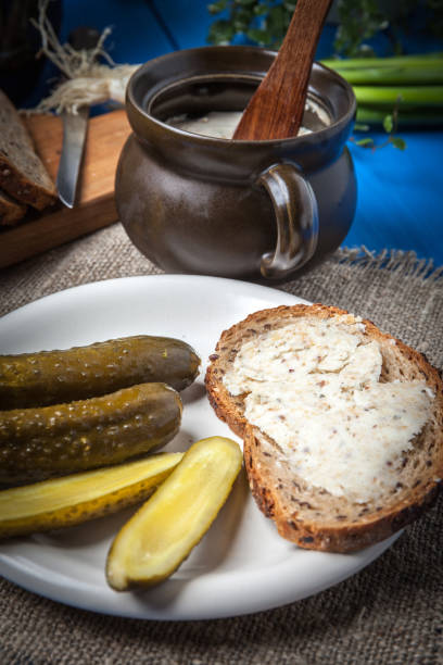 A plate of bread spread with butter, accompanied by pickles and a clay pot with a wooden spoon.