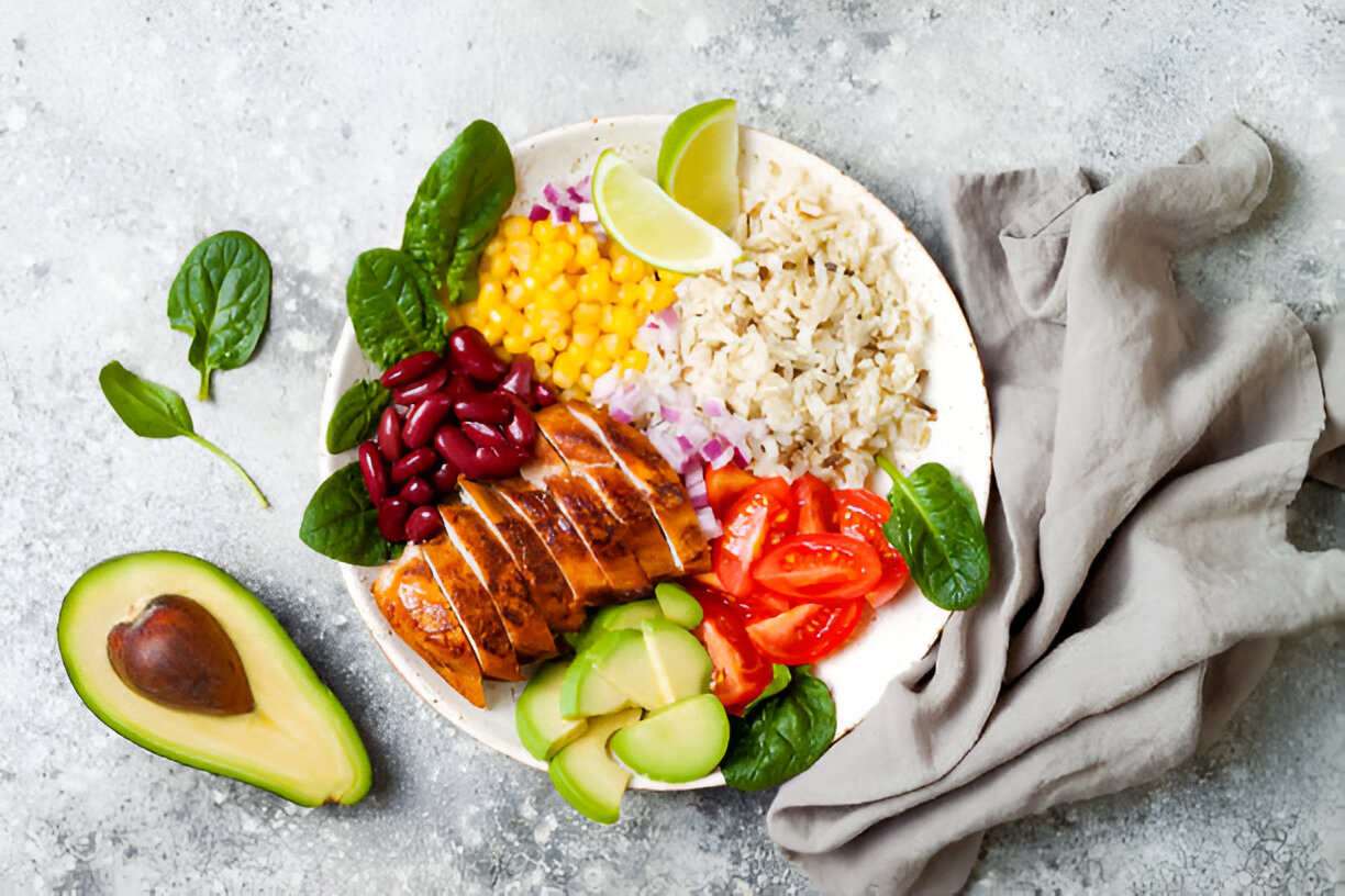 A colorful and healthy plate featuring sliced grilled chicken, corn, black beans, rice, spinach, avocado, and cherry tomatoes.