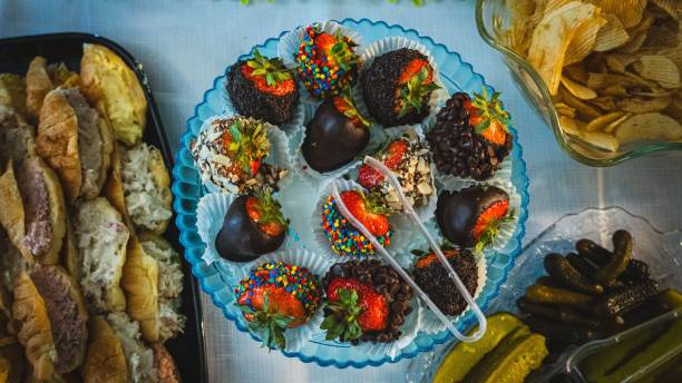 A colorful platter of chocolate-covered strawberries decorated with sprinkles and nuts, surrounded by various snacks including rolls and pickles.