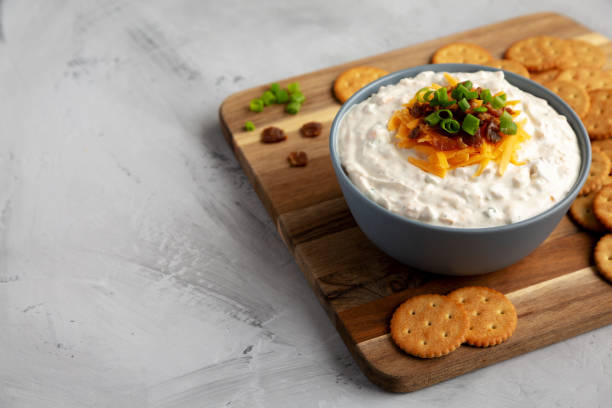 A bowl of creamy dip topped with green onions and cheese, surrounded by crackers on a wooden board.
