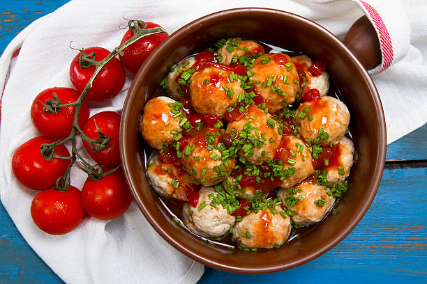 A bowl of meatballs garnished with green chives and sauce, surrounded by fresh tomatoes on a wooden table.