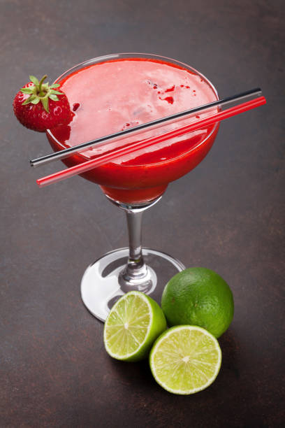 A vibrant strawberry cocktail garnished with a fresh strawberry, served with lime halves on a dark background.