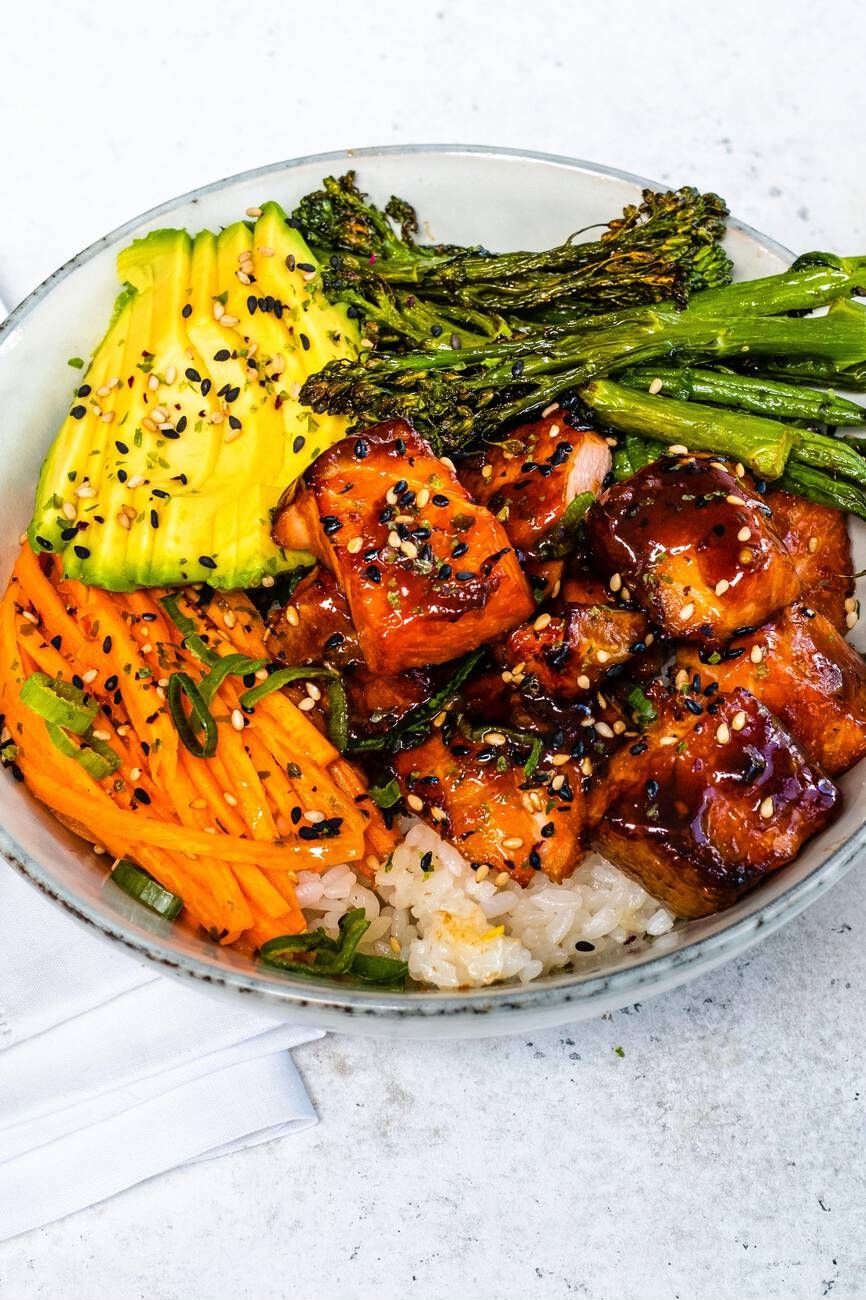 A delicious bowl of rice topped with glazed salmon, fresh vegetables including avocado, carrots, and broccoli, sprinkled with sesame seeds.