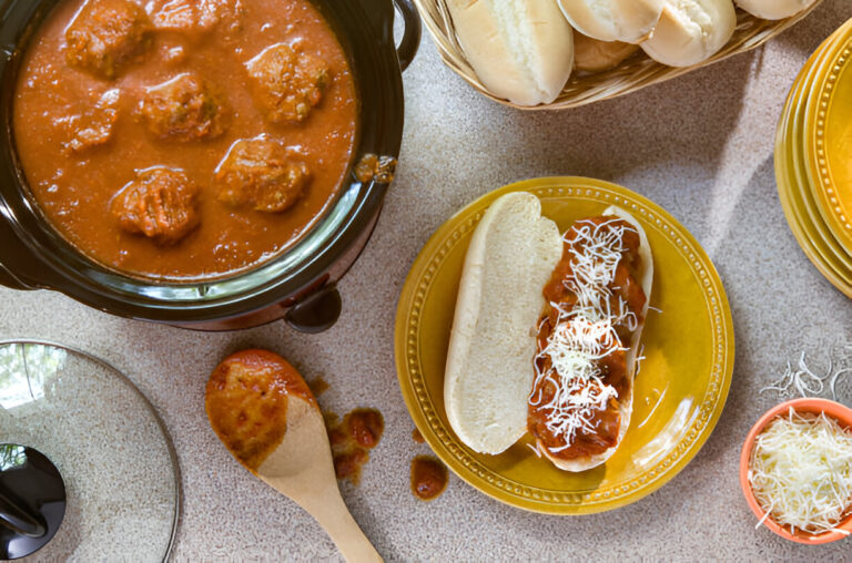 A plate with a hoagie roll topped with meatballs and marinara sauce, alongside a slow cooker of meatballs and a basket of buns.