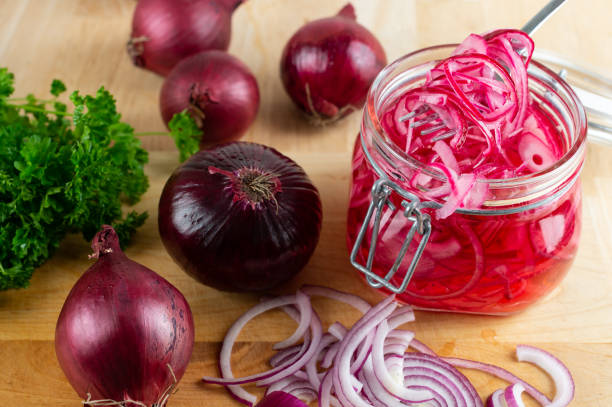 Jar of pickled red onions surrounded by whole red onions and fresh herbs on a wooden surface.
