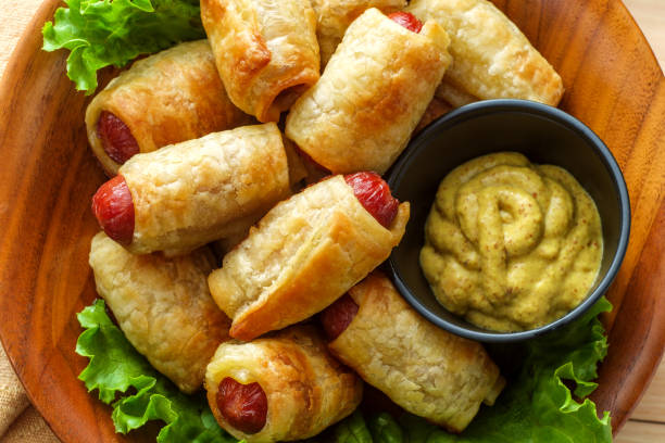 A wooden platter filled with sausage rolls resting on lettuce leaves, accompanied by a small bowl of mustard.
