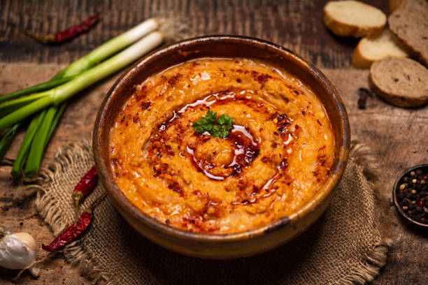A bowl of orange hummus garnished with green herbs and a drizzle of oil, surrounded by vegetables and bread.