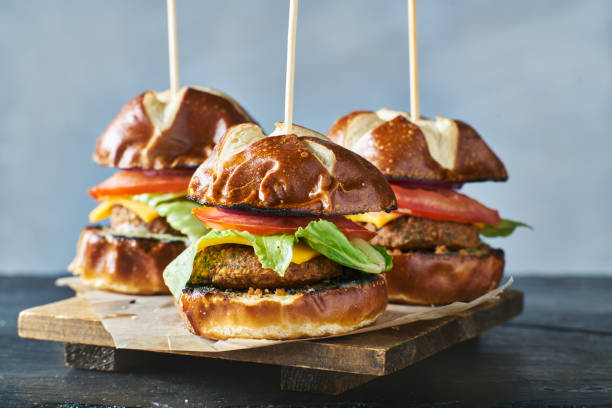 Three pretzel bun burgers with lettuce, tomato, and cheese arranged on a wooden board.
