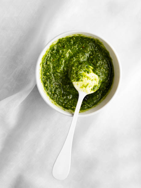 A small white bowl of green pesto with a white spoon resting inside, placed on a light gray surface.