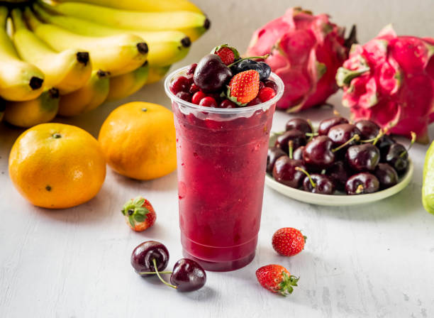 A refreshing berry drink in a clear cup surrounded by fresh fruits including bananas, oranges, and dragon fruit.