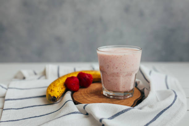 A glass of pink smoothie with banana and strawberries on a wooden board, placed on a striped towel