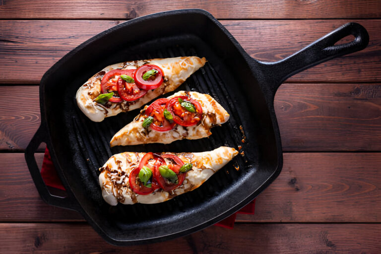 A cast iron skillet with three grilled chicken fillets topped with sliced tomatoes, red onions, and fresh basil leaves.