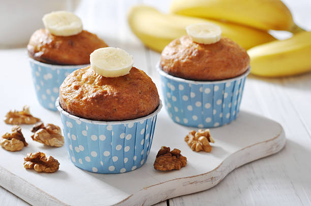 Three banana muffins topped with banana slices, in blue polka dot cups, on a wooden serving board with walnuts and bananas in the background.