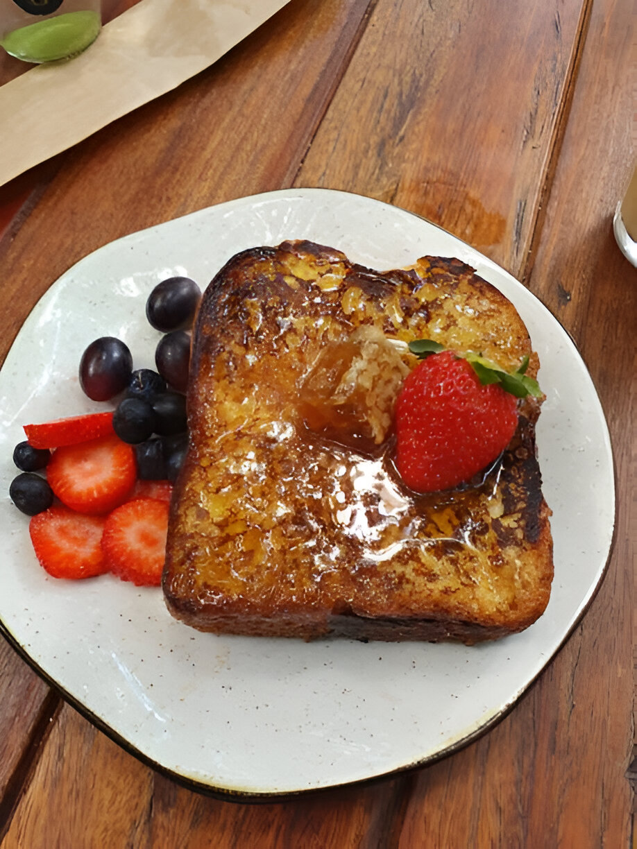 A delicious slice of French toast topped with a fresh strawberry and syrup, served with a side of mixed berries on a cream-colored plate.