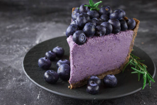 A slice of blueberry cheesecake topped with fresh blueberries on a dark plate, with additional blueberries scattered around.