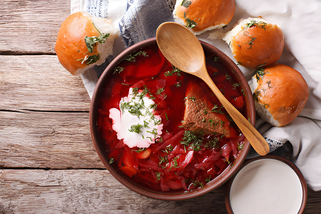 A bowl of vibrant red borscht with a dollop of sour cream, garnished with herbs, alongside soft bread rolls on a wooden surface.