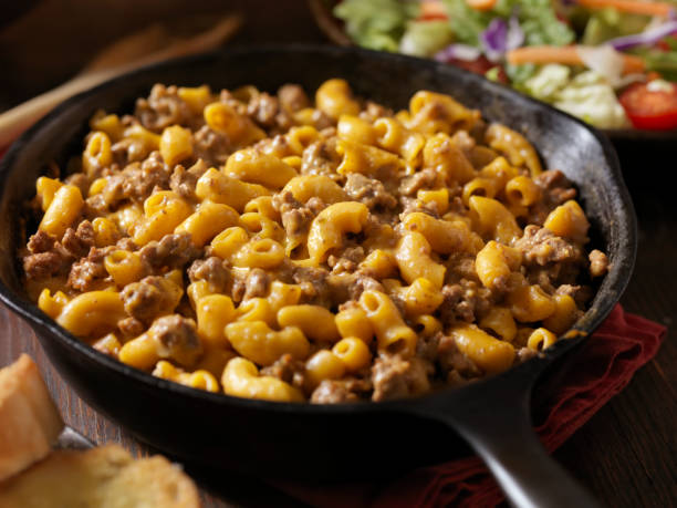 A skillet filled with cheesy macaroni and beef, with a side of salad and bread in the background.