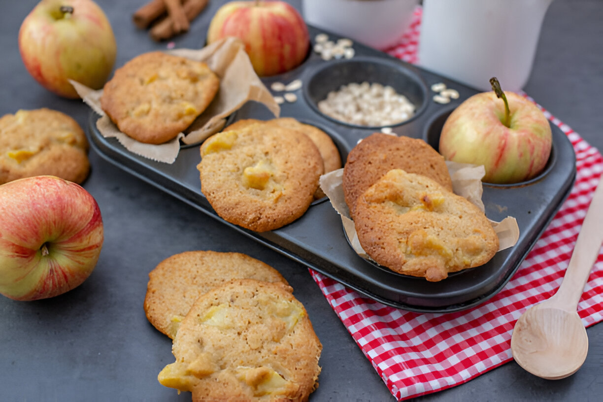 Freshly baked apple cookies on a baking tray with whole apples and a wooden spoon
