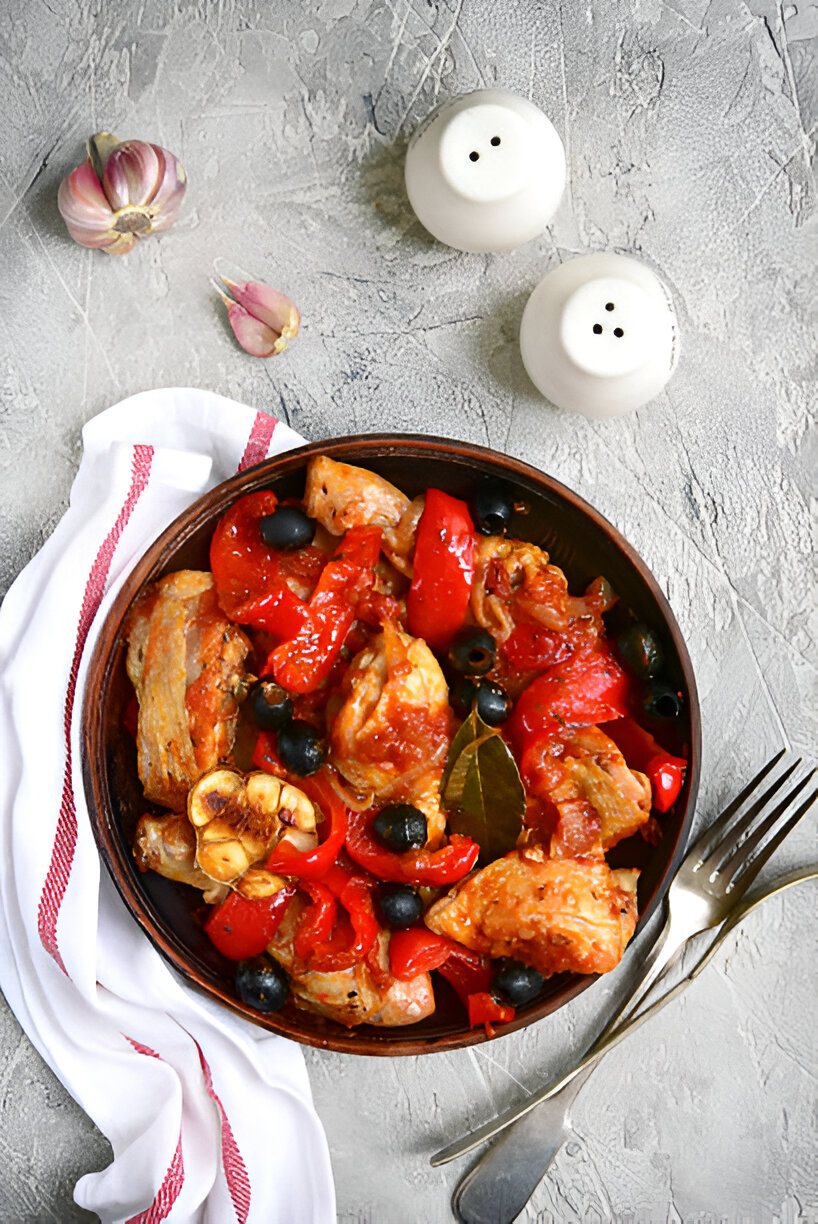 A rustic dish of chicken, red peppers, and olives served in a brown bowl on a textured surface.
