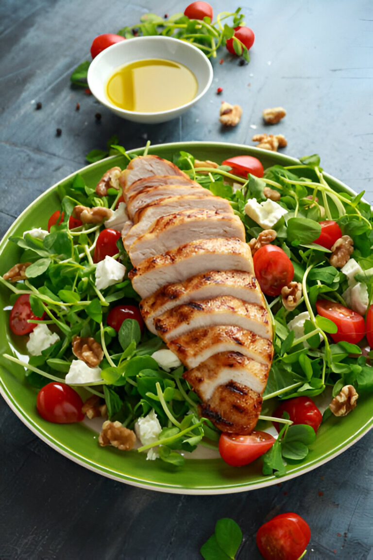 Grilled chicken salad featuring sliced grilled chicken breast, cherry tomatoes, walnuts, and feta cheese on a bed of mixed greens, with olive oil in a small bowl in the background.