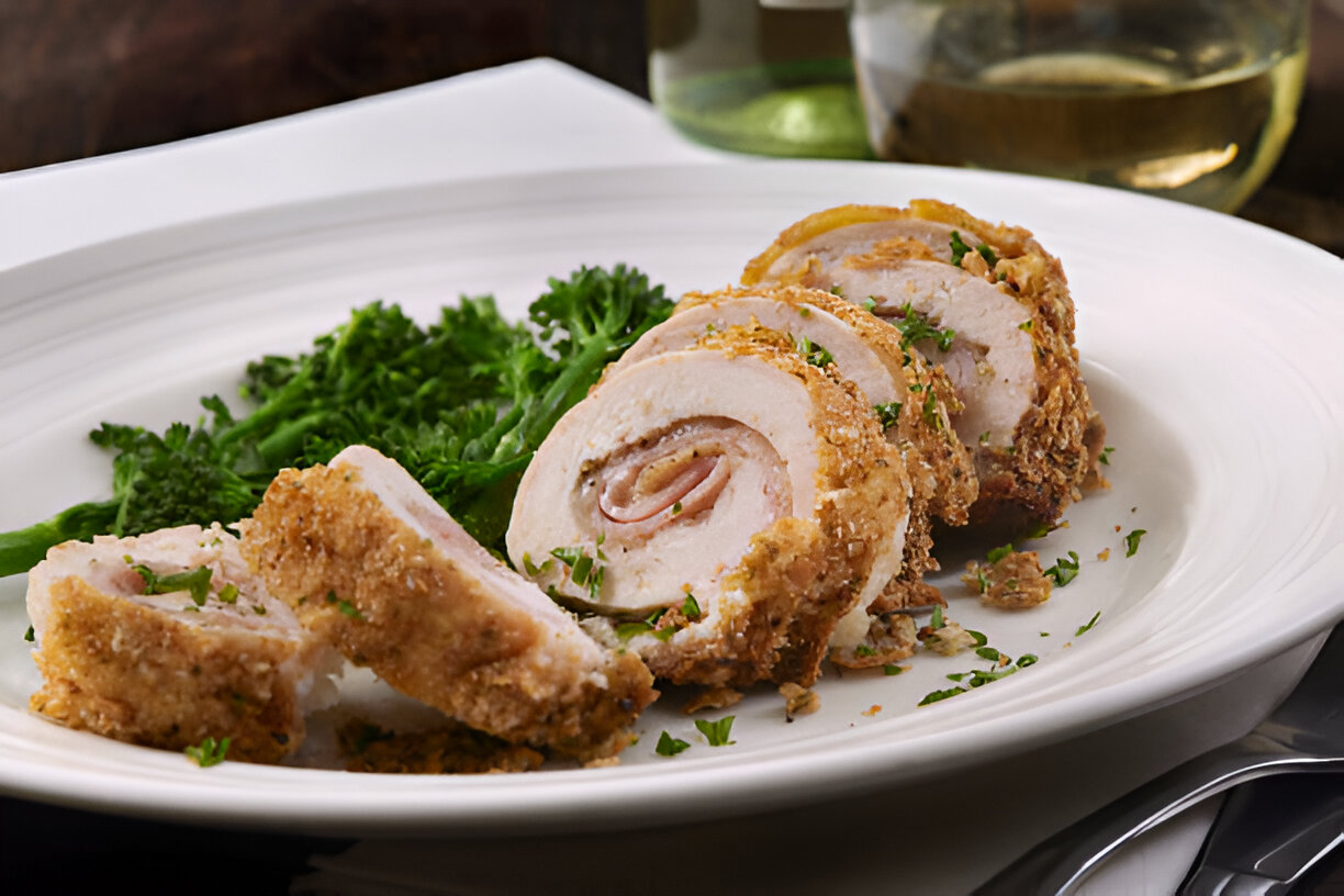 Slices of stuffed chicken rolled with ham, garnished with parsley and served with greens on a white plate.