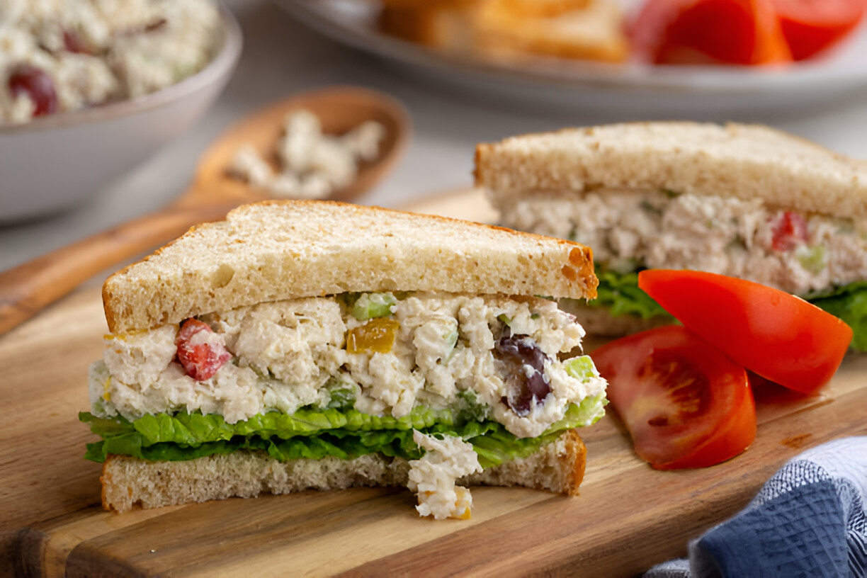 A close-up of a sandwich filled with chicken salad, lettuce, and served with slices of tomato on a wooden cutting board.